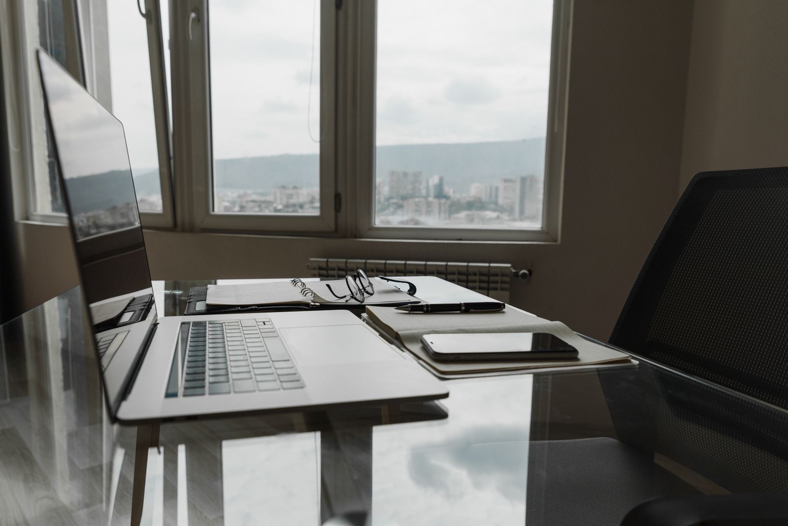 A laptop and an open notebook on a desk by a tall window